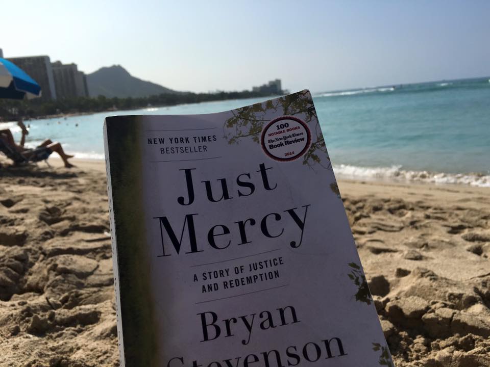 Picture of the cover of the book Just Mercy written by Bryan Stevenson, being held with the backdrop of Waikiki beach.